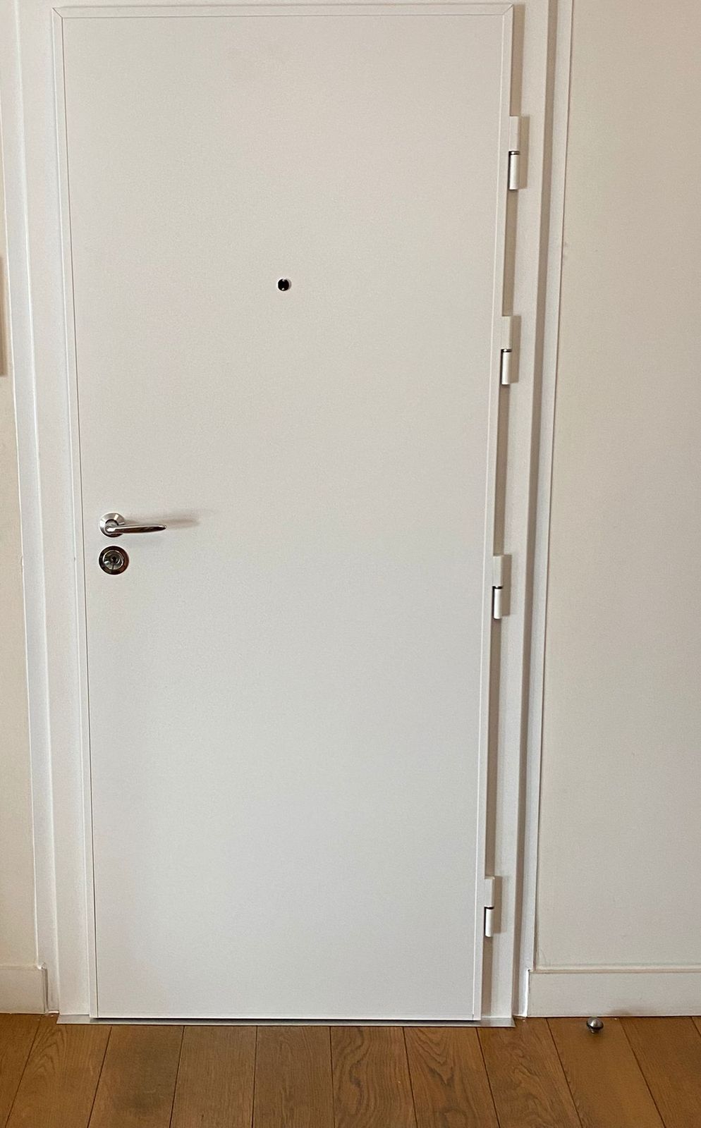 Porte blindée blanche lisse aux paumelles apparentes, dans une entrée au parquet en chêne