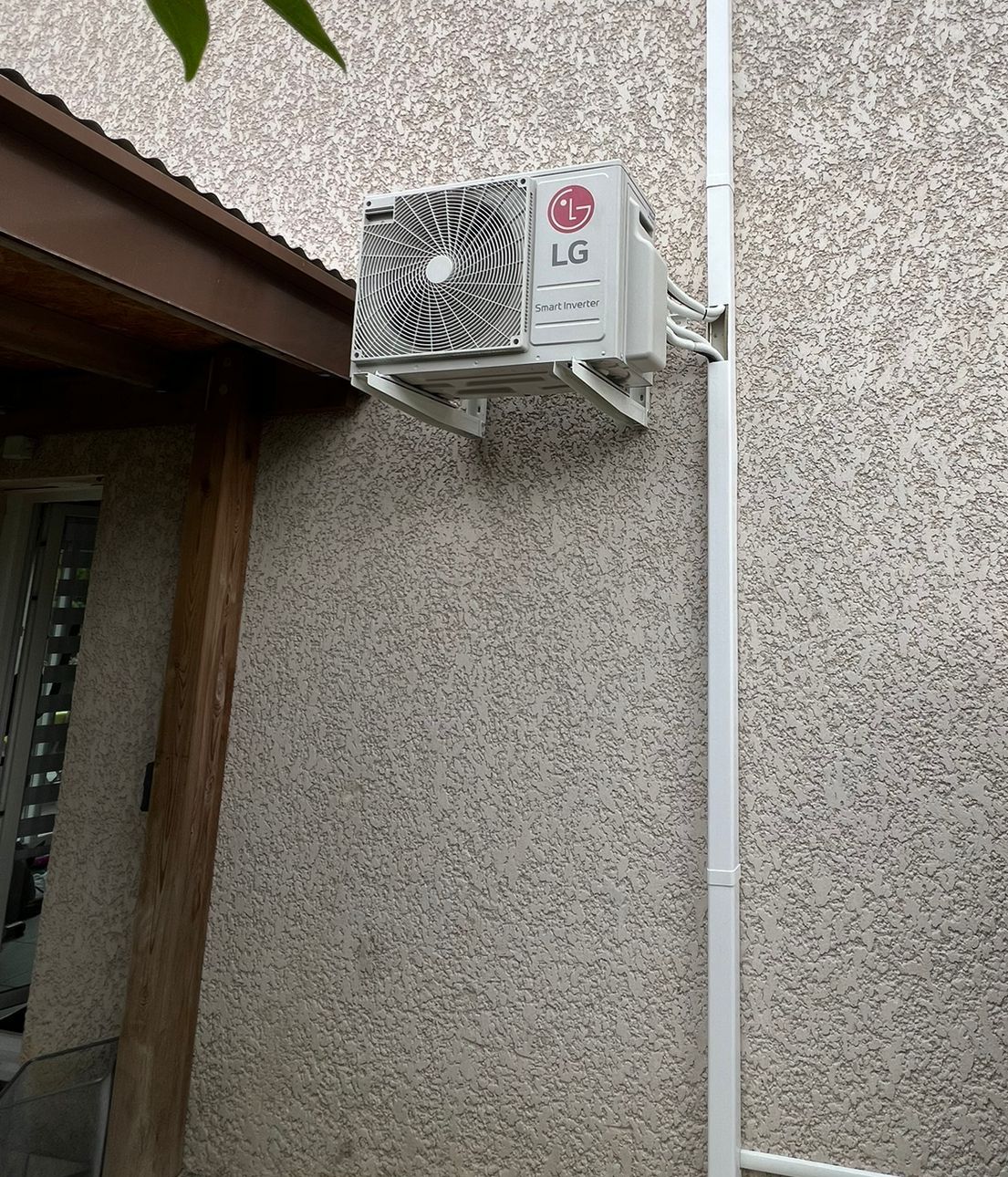 Unité extérieure de climatisation fixée en hauteur sur un mur crépi, avec goulotte blanche