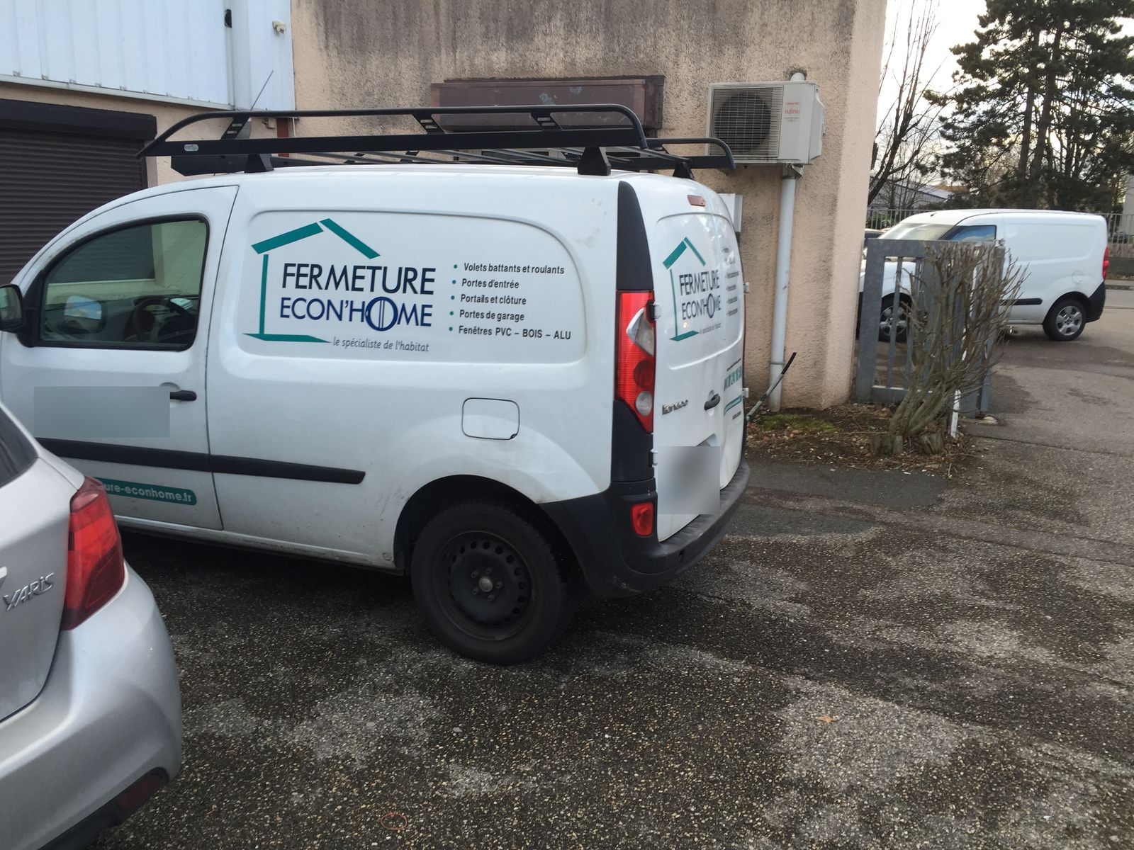 Véhicule utilitaire blanc floqué Fermeture Econ'Home, avec galerie de toit, stationné devant un bâtiment