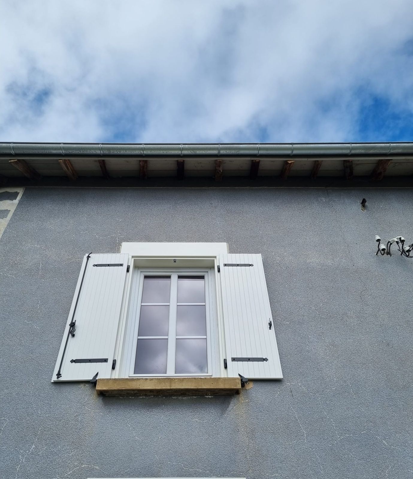Fenêtre blanche à petits bois et volets battants blancs sur une façade grise, ciel nuageux