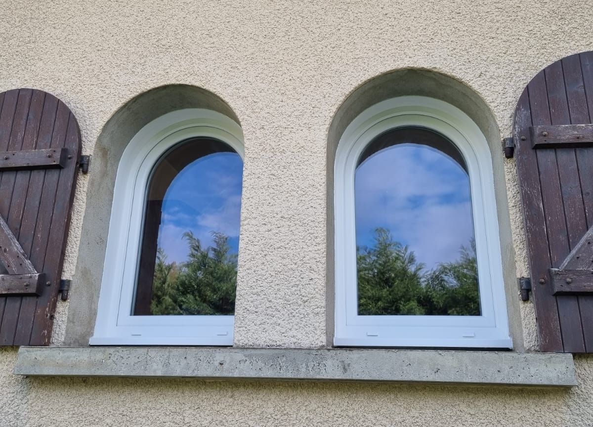 Deux fenêtres cintrées blanches posées dans des baies en arc, entre des volets battants en bois