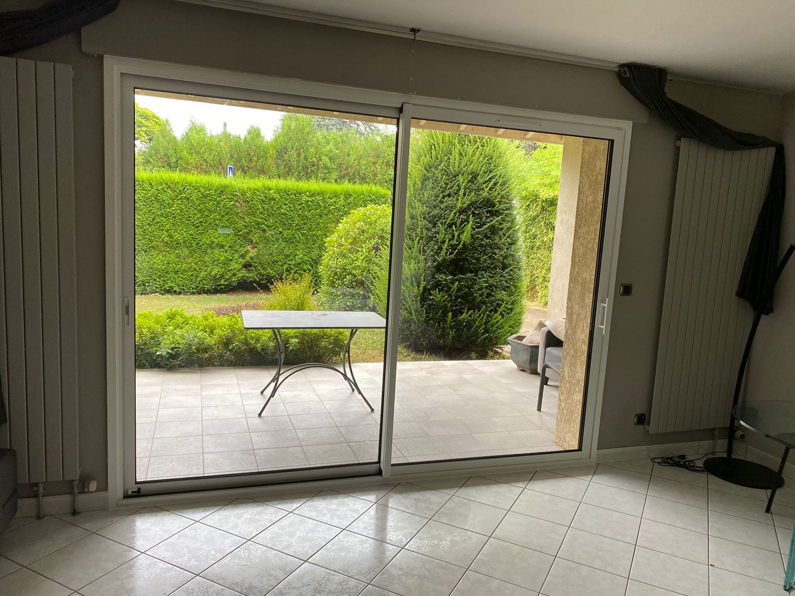 Baie vitrée coulissante blanche donnant sur une terrasse carrelée et un jardin entouré de haies