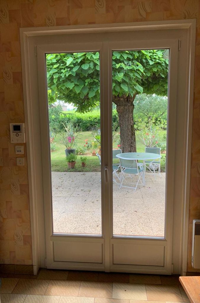 Porte-fenêtre blanche à deux vantaux ouvrant sur une terrasse ombragée par un arbre