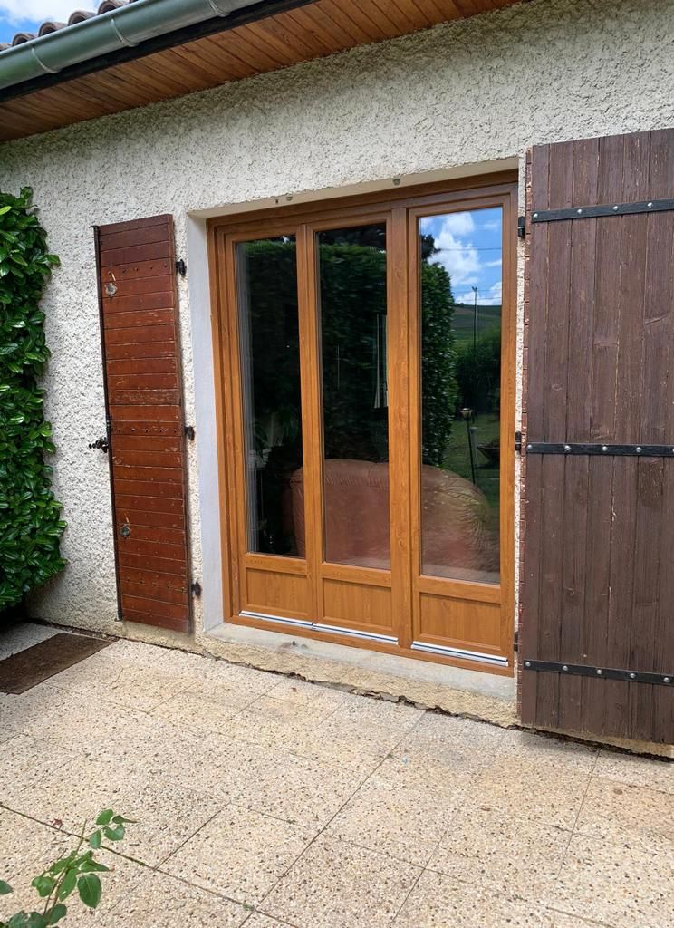 Porte-fenêtre à trois vantaux ton chêne doré, encadrée de volets battants en bois, sur une façade crépie