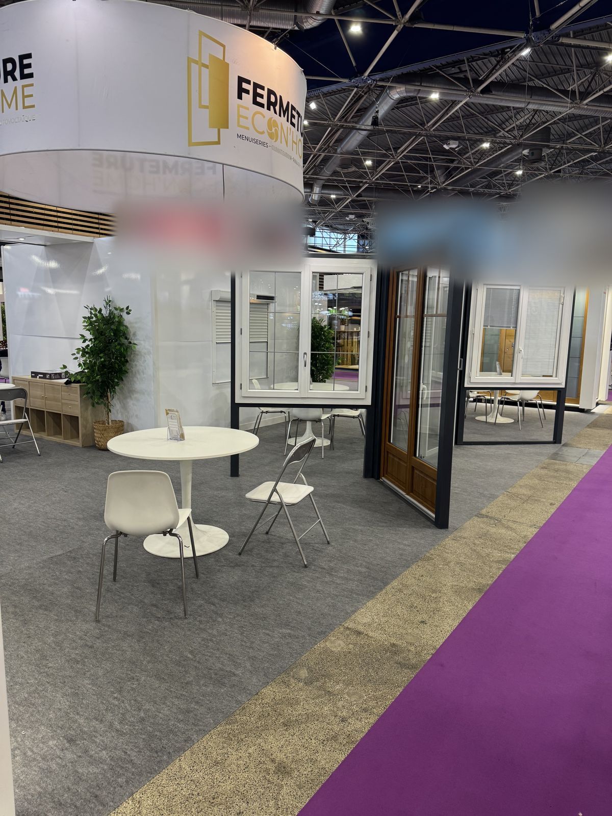Vue d'ensemble du stand à Eurexpo : coin salon avec tables blanches, fenêtres et porte-fenêtre ton bois sous l'enseigne ronde Fermeture Econ'Home