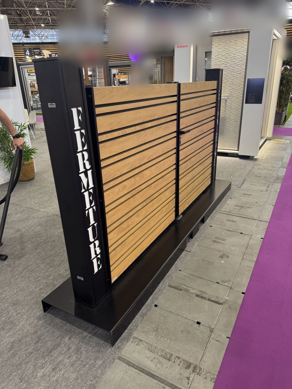 Clôture en aluminium ton bois exposée sur le stand, avec le nom Fermeture sur son poteau