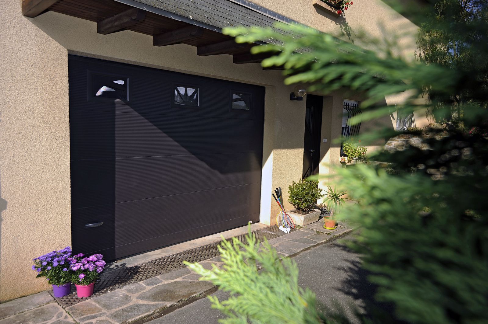 Porte de garage sectionnelle gris anthracite avec une rangée de hublots rectangulaires, devant une allée fleurie