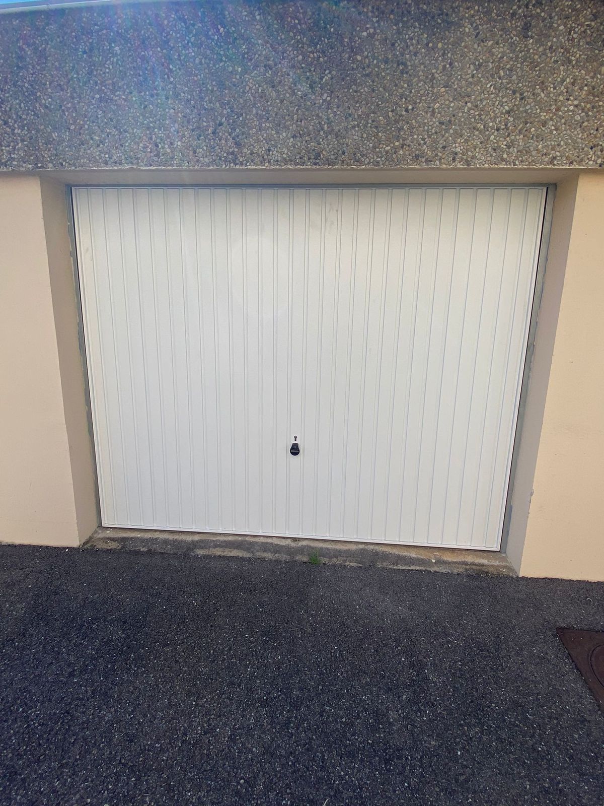 Porte de garage basculante blanche à rainures verticales, devant une allée en enrobé
