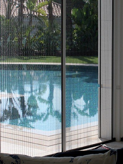 Moustiquaire plissée PLISSEO à toile noire tirée devant une baie vitrée, avec vue sur une piscine et des palmiers