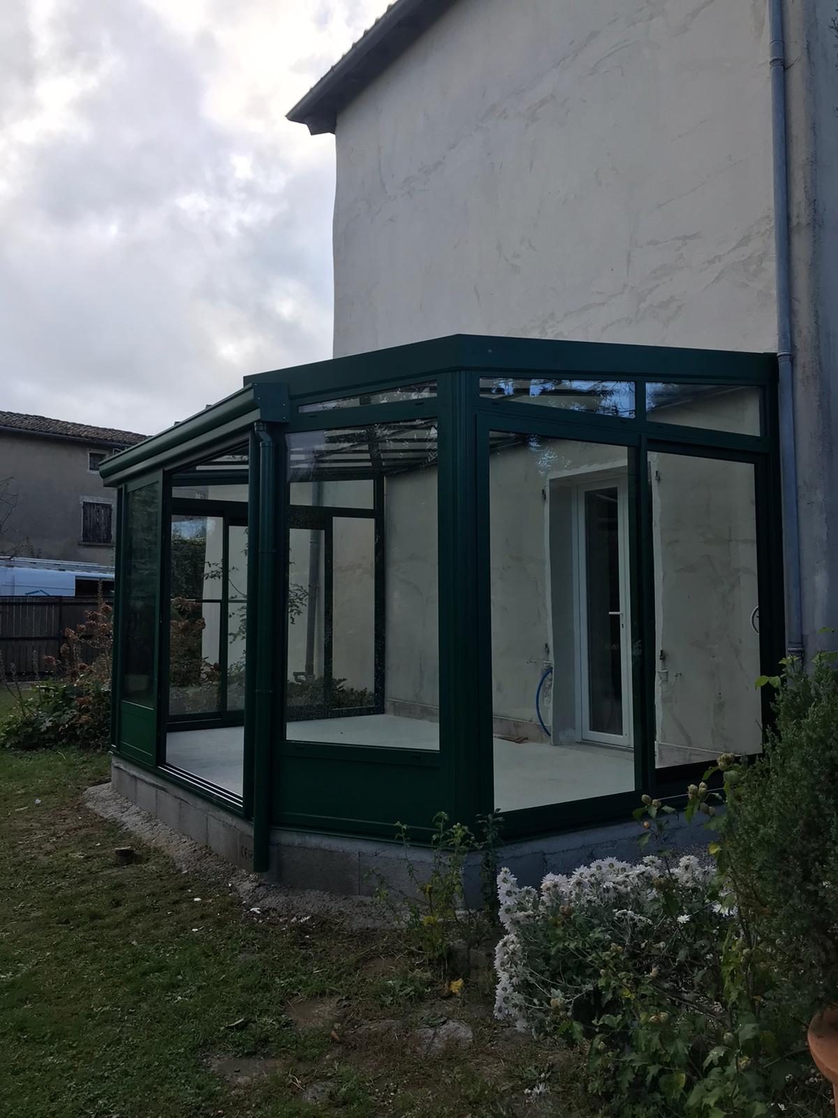 Véranda à profilés verts adossée à une maison crépie, vue de côté depuis le jardin