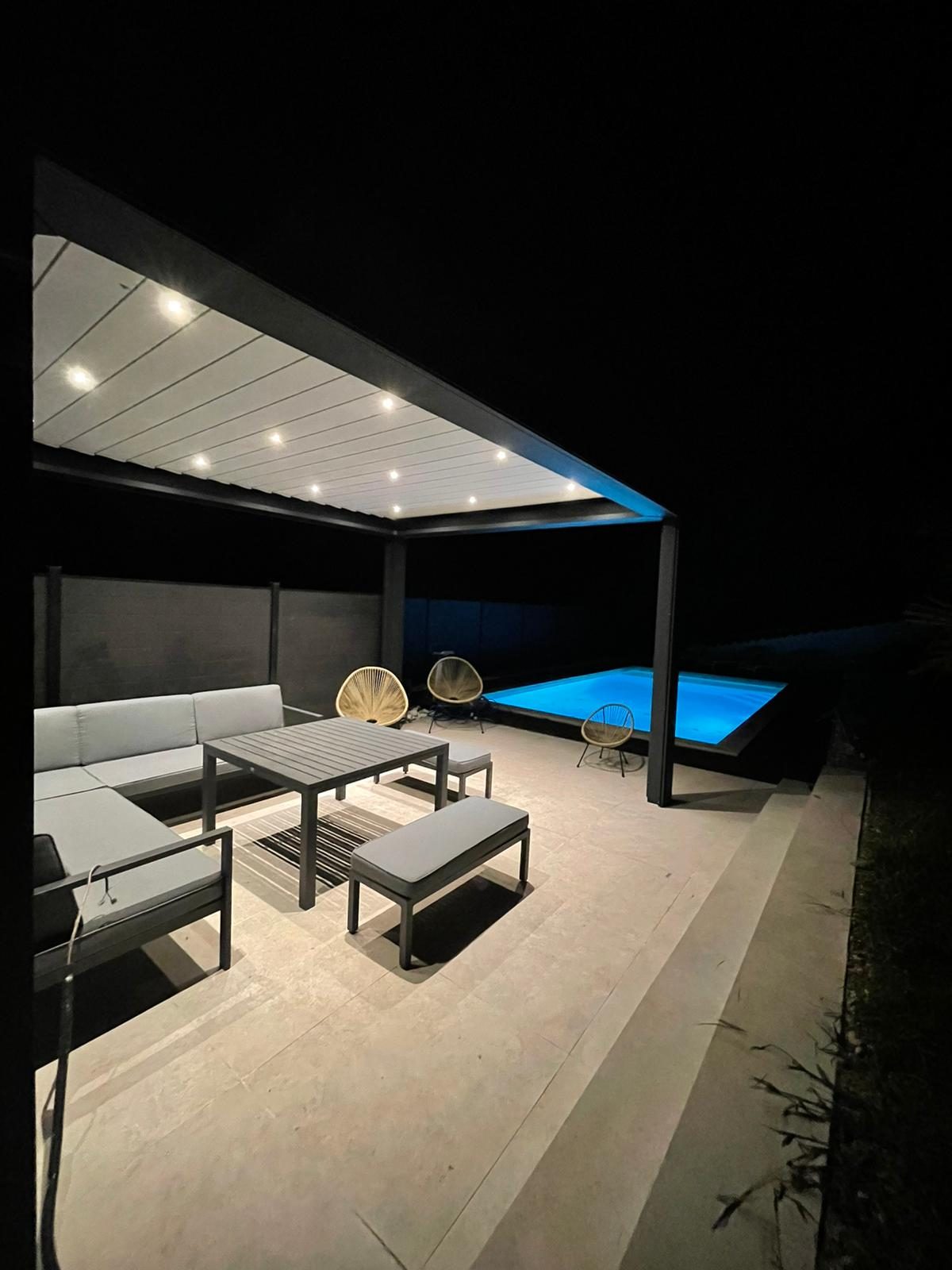 Pergola à lames gris anthracite éclairée de nuit par des spots intégrés, au-dessus d'un salon de jardin près d'une piscine