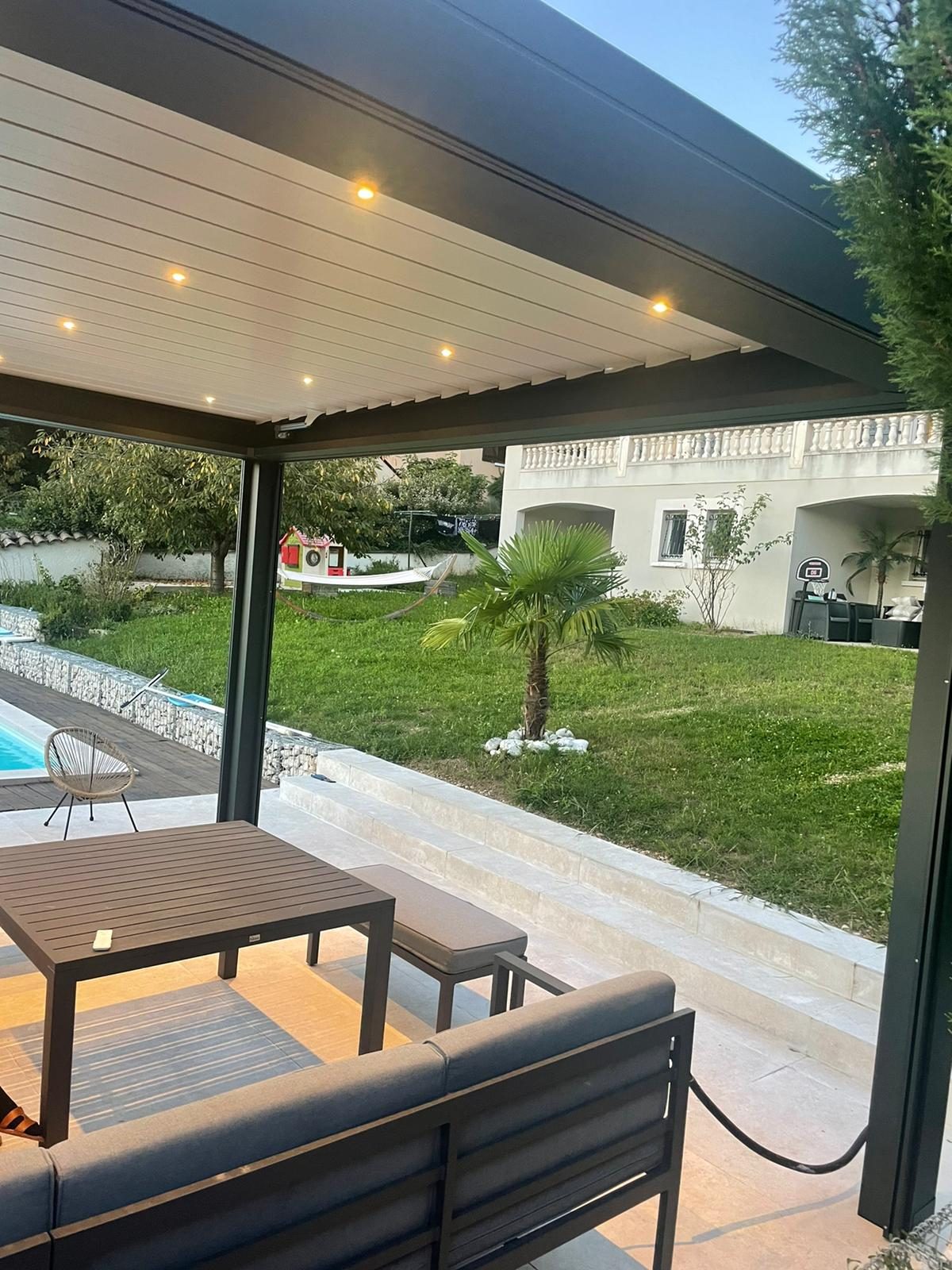 Pergola à lames gris anthracite avec spots, abritant une table de jardin face à la pelouse et à la piscine