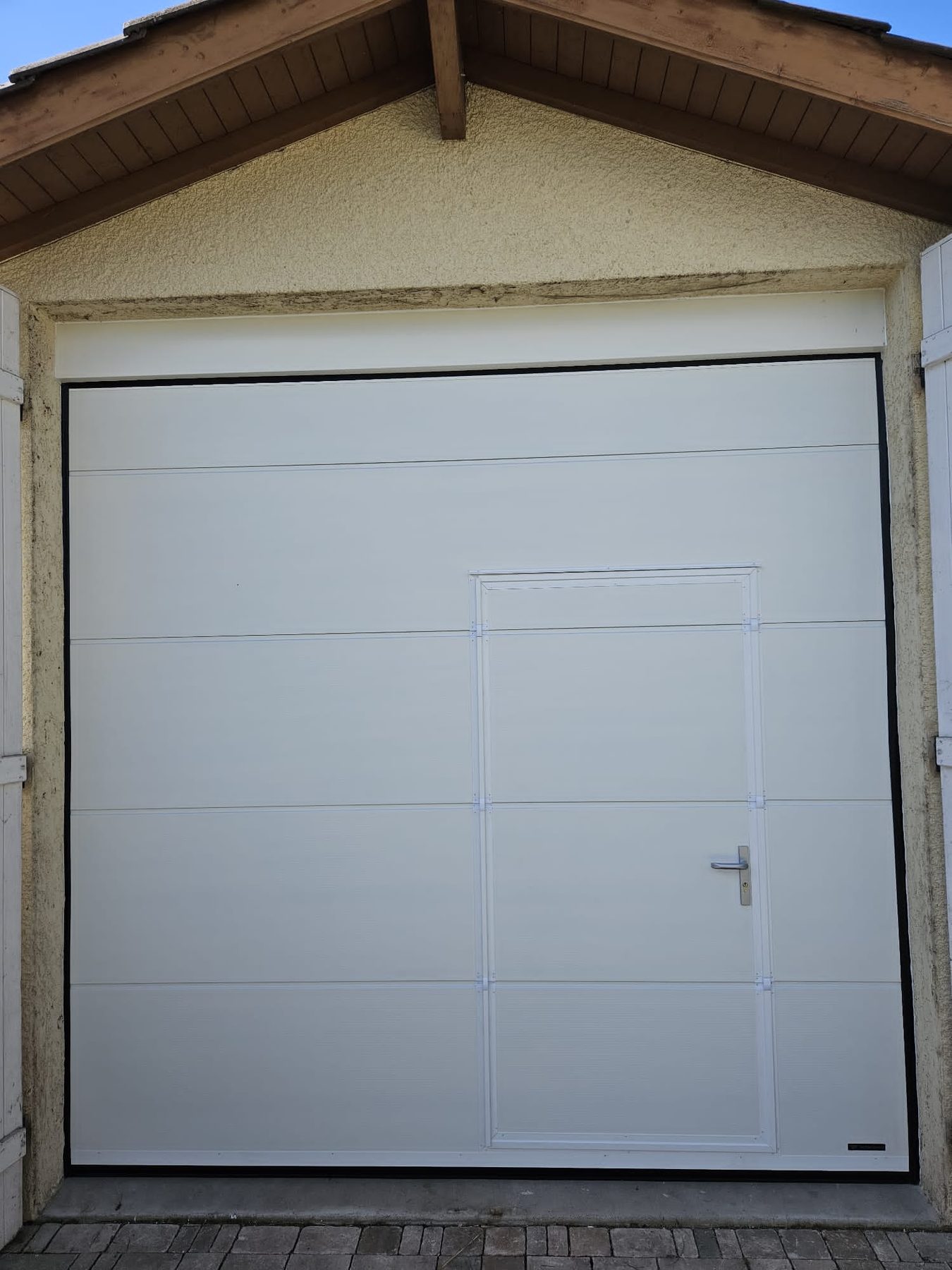 Porte de garage sectionnelle blanche avec portillon intégré, sous un avant-toit en bois