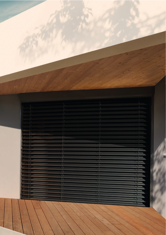 Brise-soleil orientable à lames gris anthracite fermé devant une grande baie, sous un avant-toit en bois, terrasse en lames