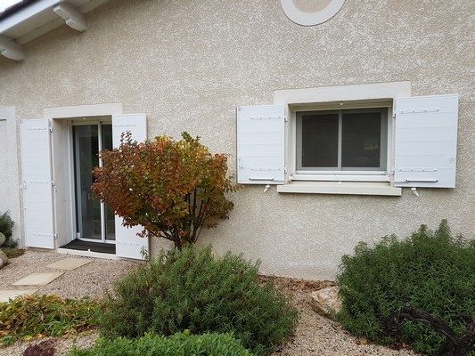 Volets battants en aluminium blanc ouverts sur la façade crépie d'une maison, jardin arboré