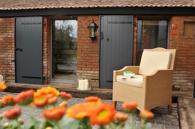 Volets battants PVC gris anthracite ouverts sur une façade en briques, fauteuil en osier et fleurs orange au premier plan