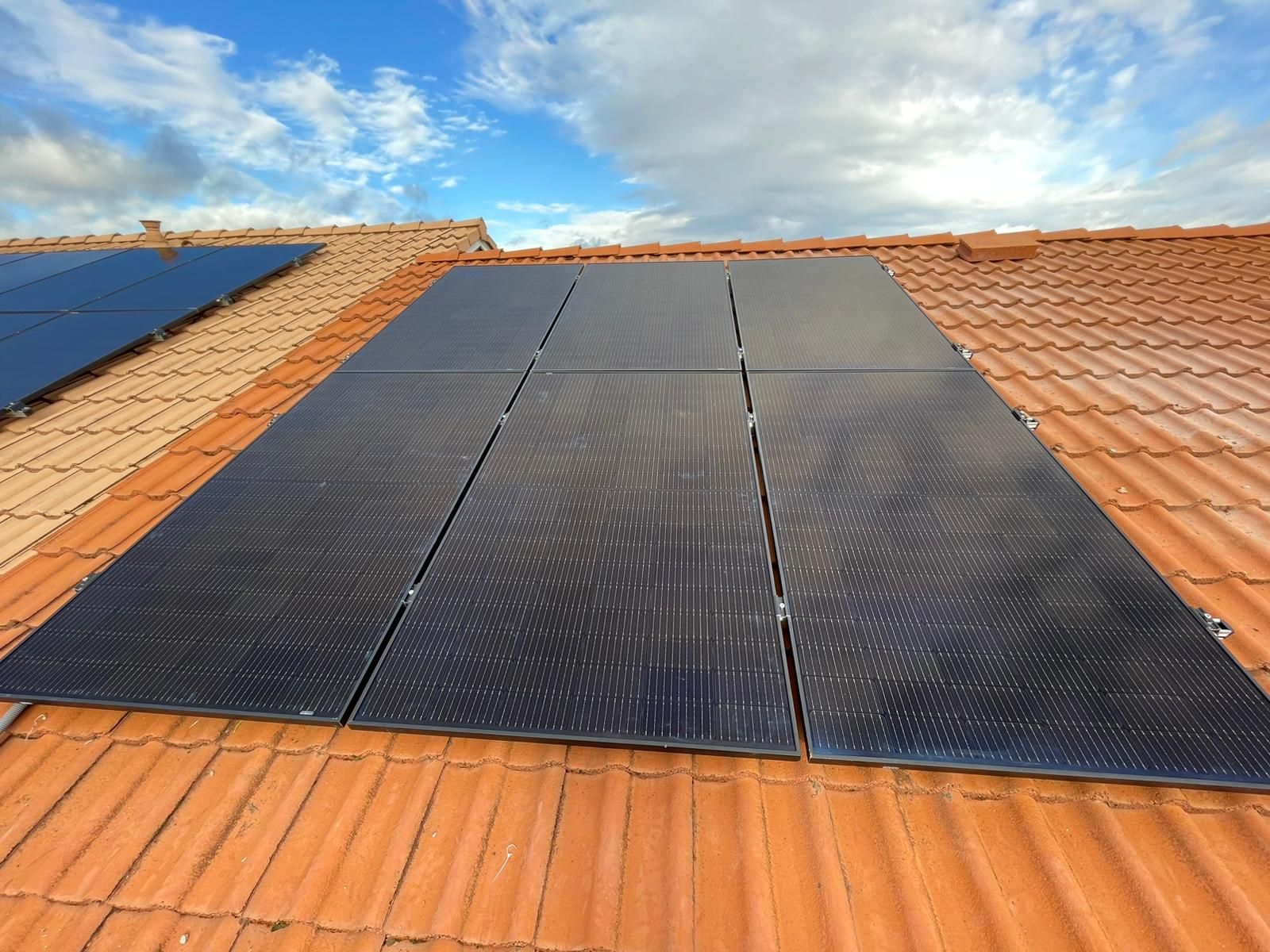 Six panneaux photovoltaïques noirs alignés sur un toit en tuiles orange, ciel nuageux