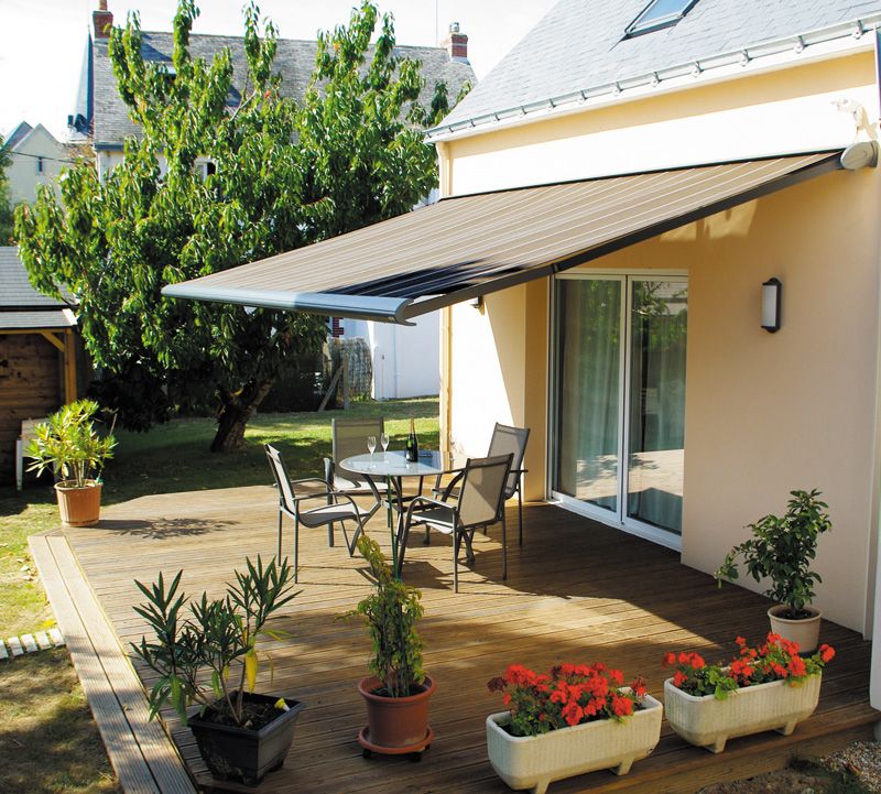 Store banne à toile grise déployé au-dessus de la terrasse en bois d'une maison, avec table et chaises de jardin