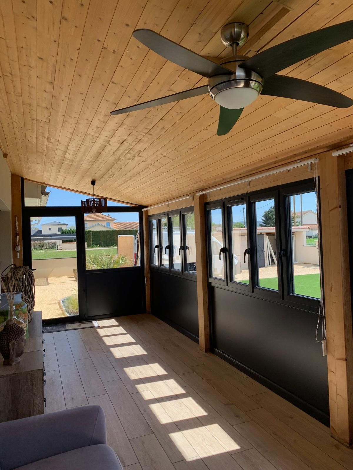 Intérieur d'une véranda lumineuse avec plafond en bois, menuiseries noires et ventilateur de plafond
