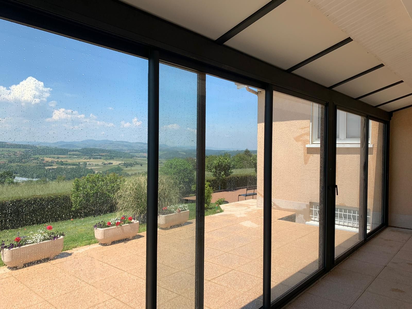 Intérieur d'une véranda aux grandes baies vitrées coulissantes, avec vue sur une terrasse et un panorama de collines