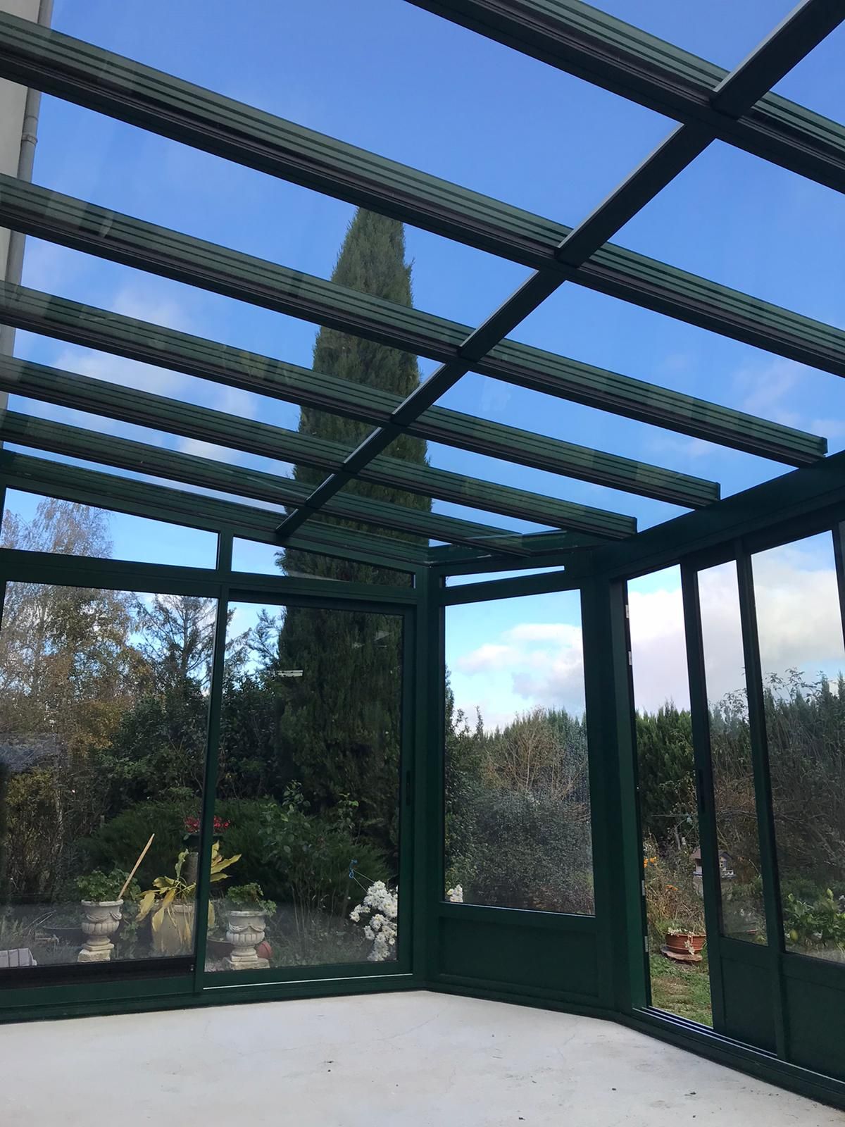 Intérieur d'une véranda aux profilés verts avec toiture vitrée, ouverte sur le jardin et le ciel