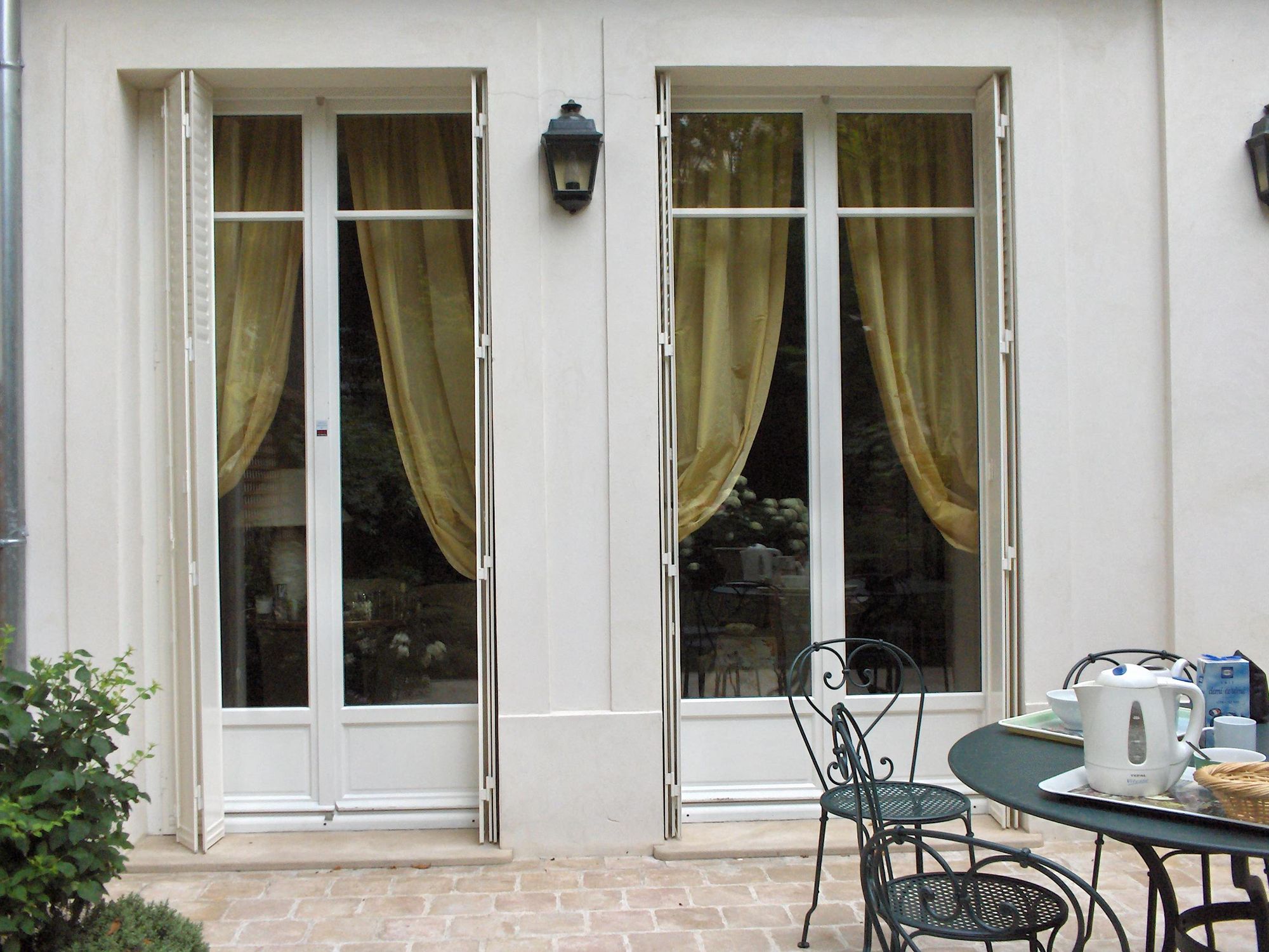 Deux portes-fenêtres d'une maison bourgeoise avec persiennes blanches repliées en tableau, terrasse avec table en fer forgé