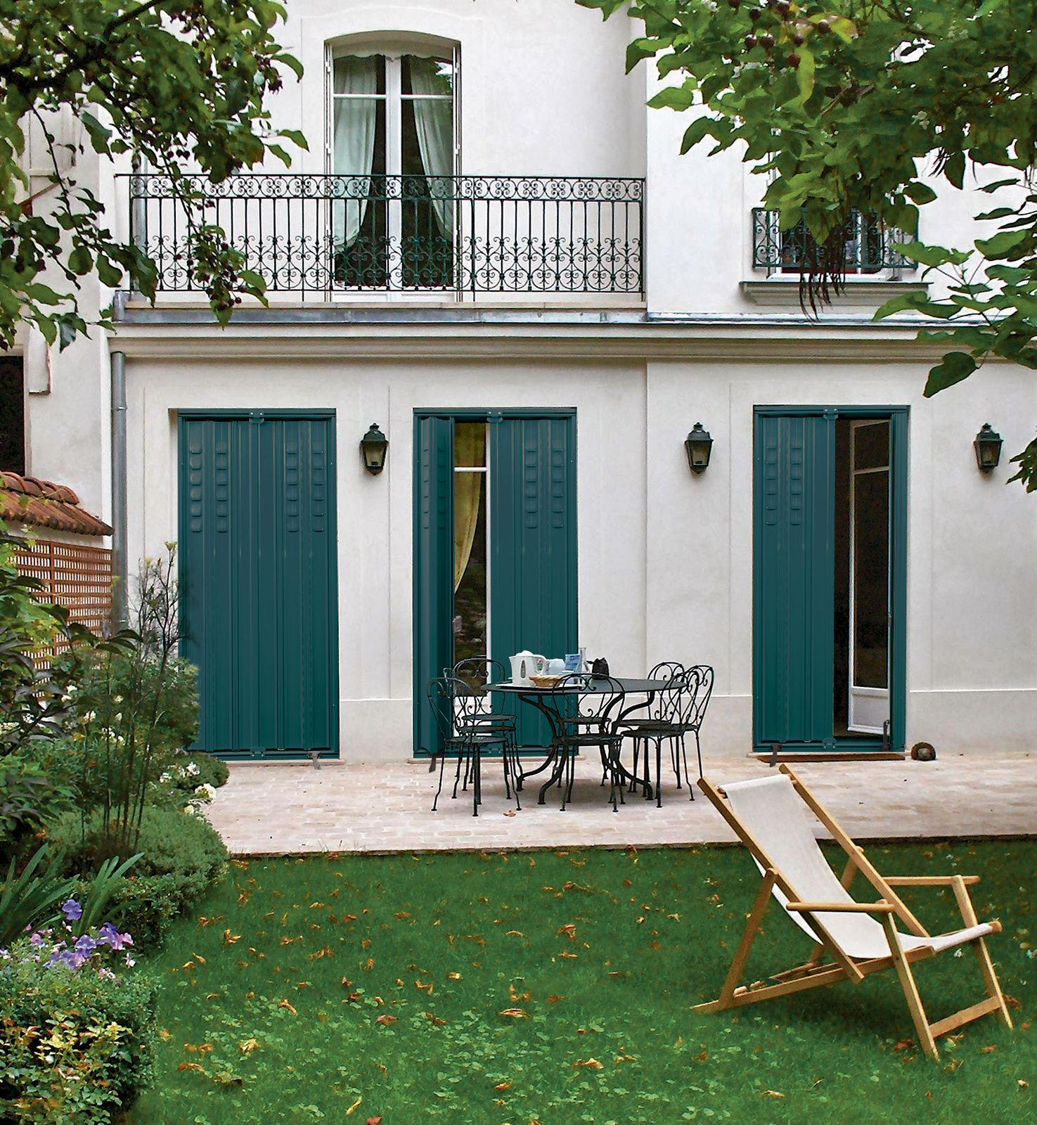 Façade de maison de maître avec persiennes vert foncé sur les portes-fenêtres du rez-de-jardin, pelouse et transat
