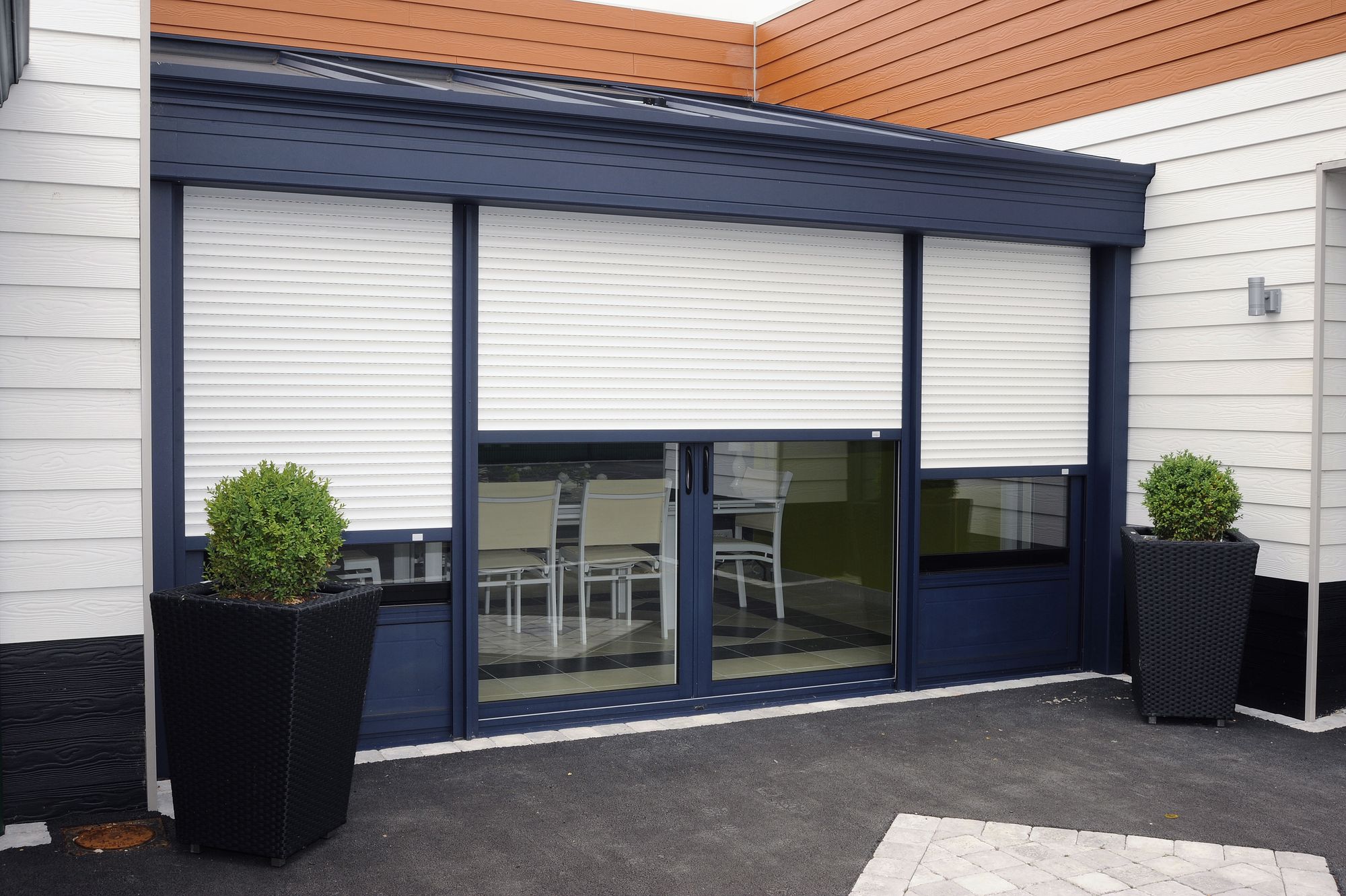 Maison contemporaine aux grandes baies vitrées gris anthracite équipées de volets roulants blancs, terrasse et bacs de buis