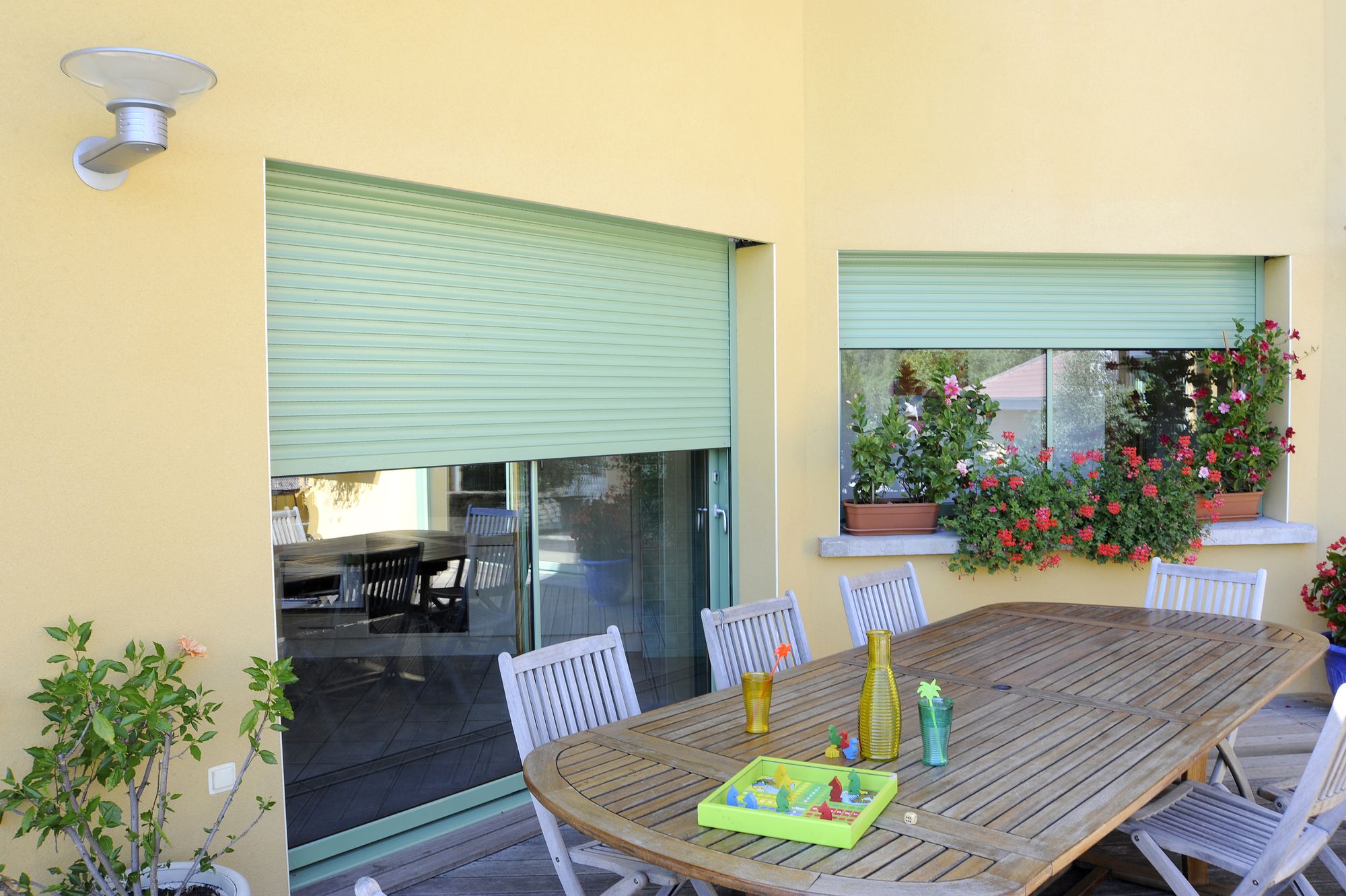 Volets roulants vert céladon sur une baie et une fenêtre de façade jaune, terrasse avec table en bois et géraniums