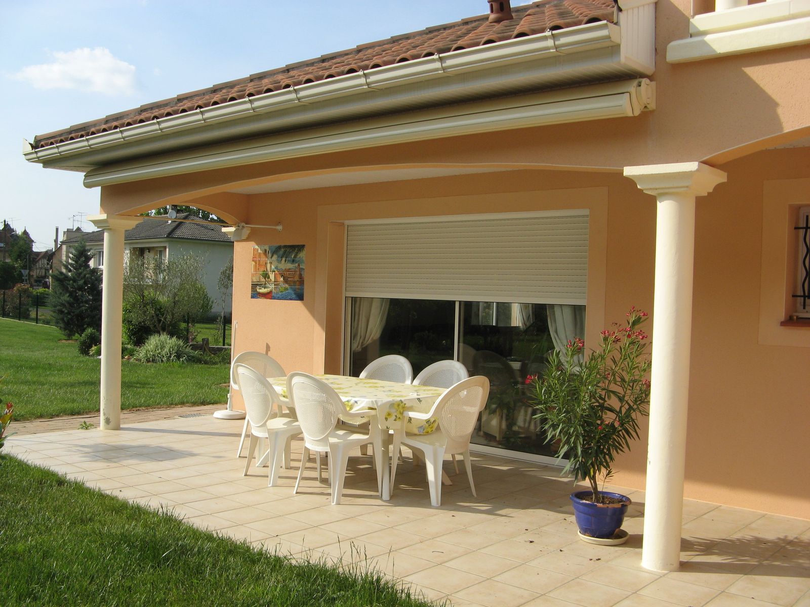 Terrasse couverte d'une maison crépie avec baie vitrée équipée d'un volet roulant et salon de jardin blanc