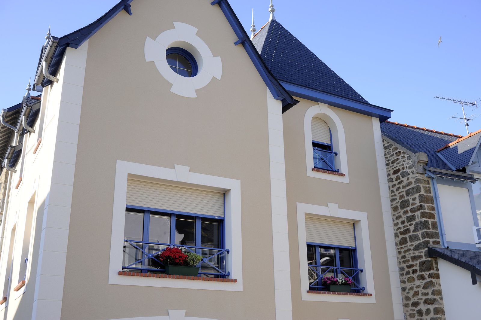 Façade de maison de ville aux fenêtres à menuiseries bleues équipées de volets roulants beiges à demi baissés, jardinières fleuries