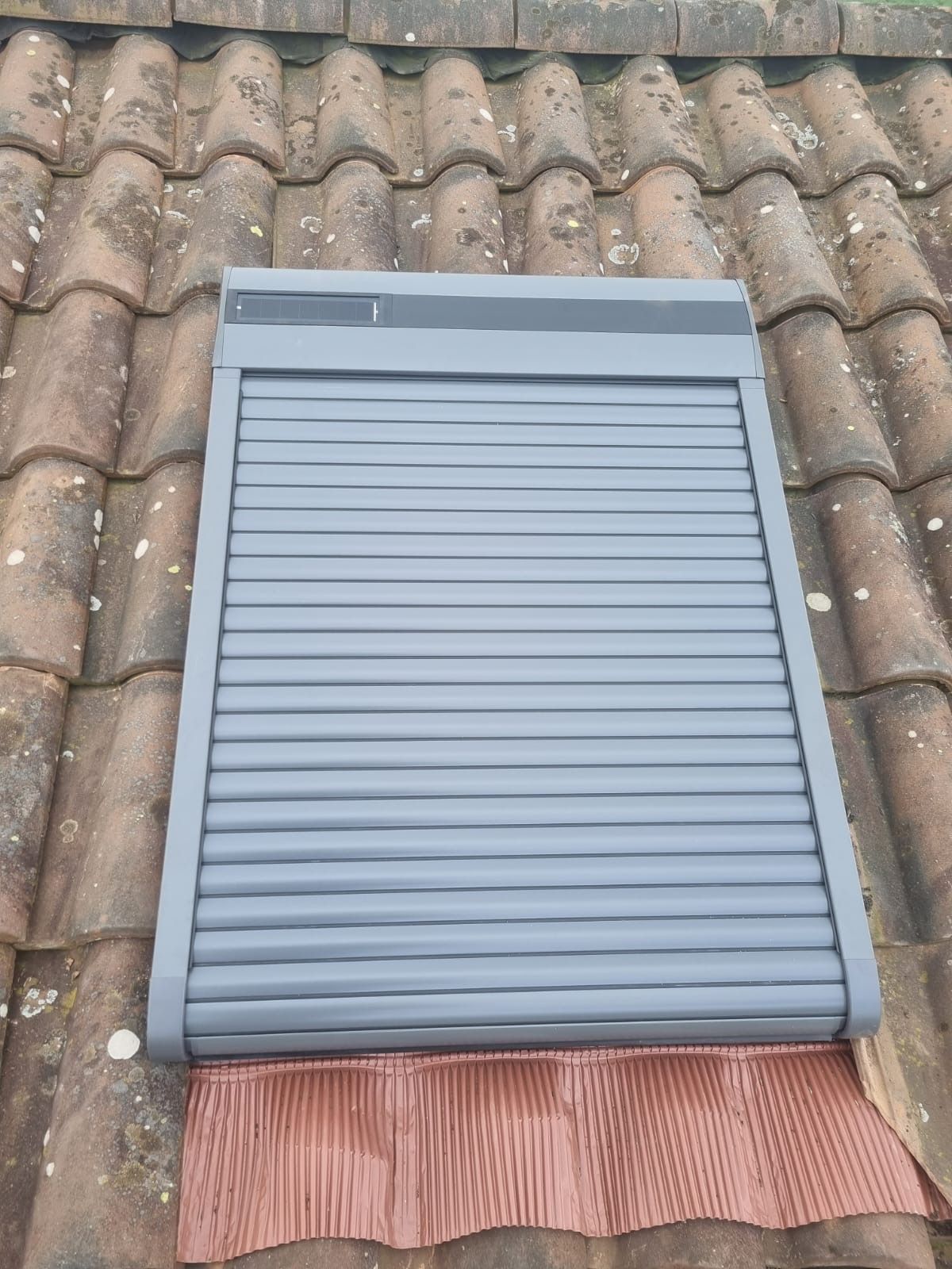 Volet roulant gris installé sur une fenêtre de toit au milieu des tuiles