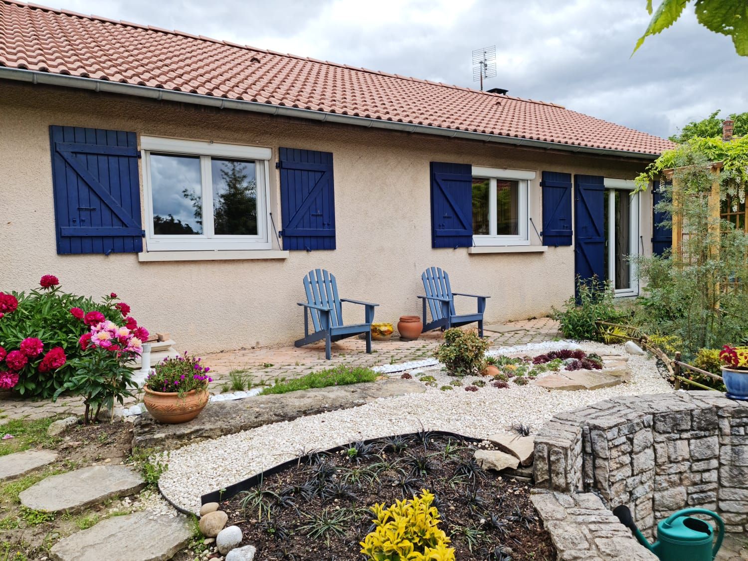 Maison de plain-pied aux fenêtres blanches et volets battants bleus, devant un jardin fleuri