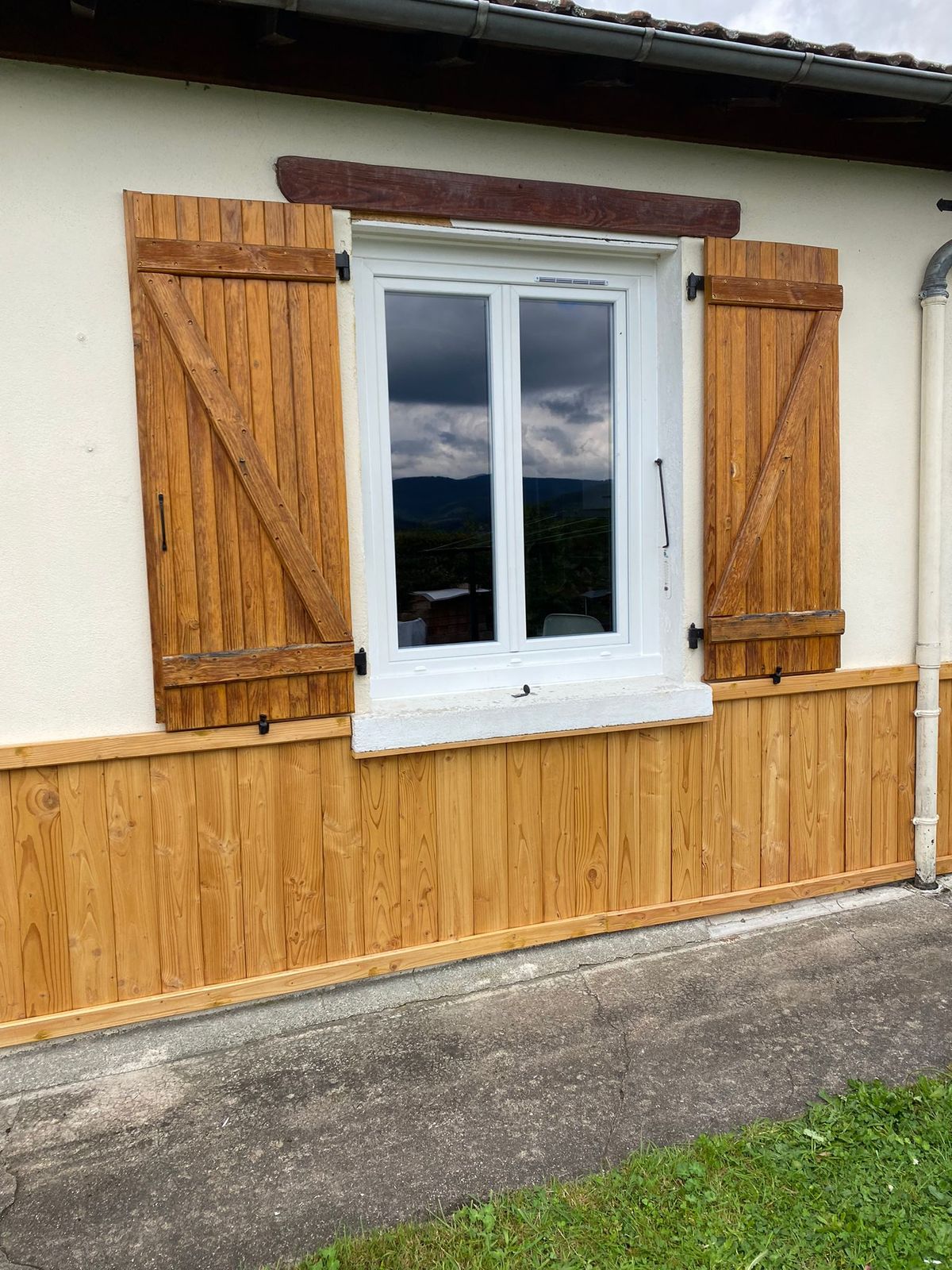 Fenêtre blanche encadrée de volets battants en bois naturel à barres et écharpe